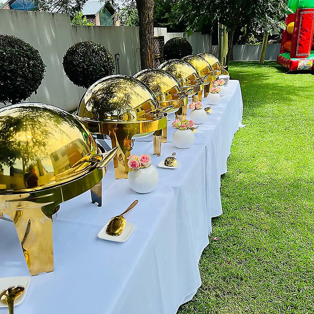 Round gold buffet chafing dishes arranged on a white-clothed outdoor event table